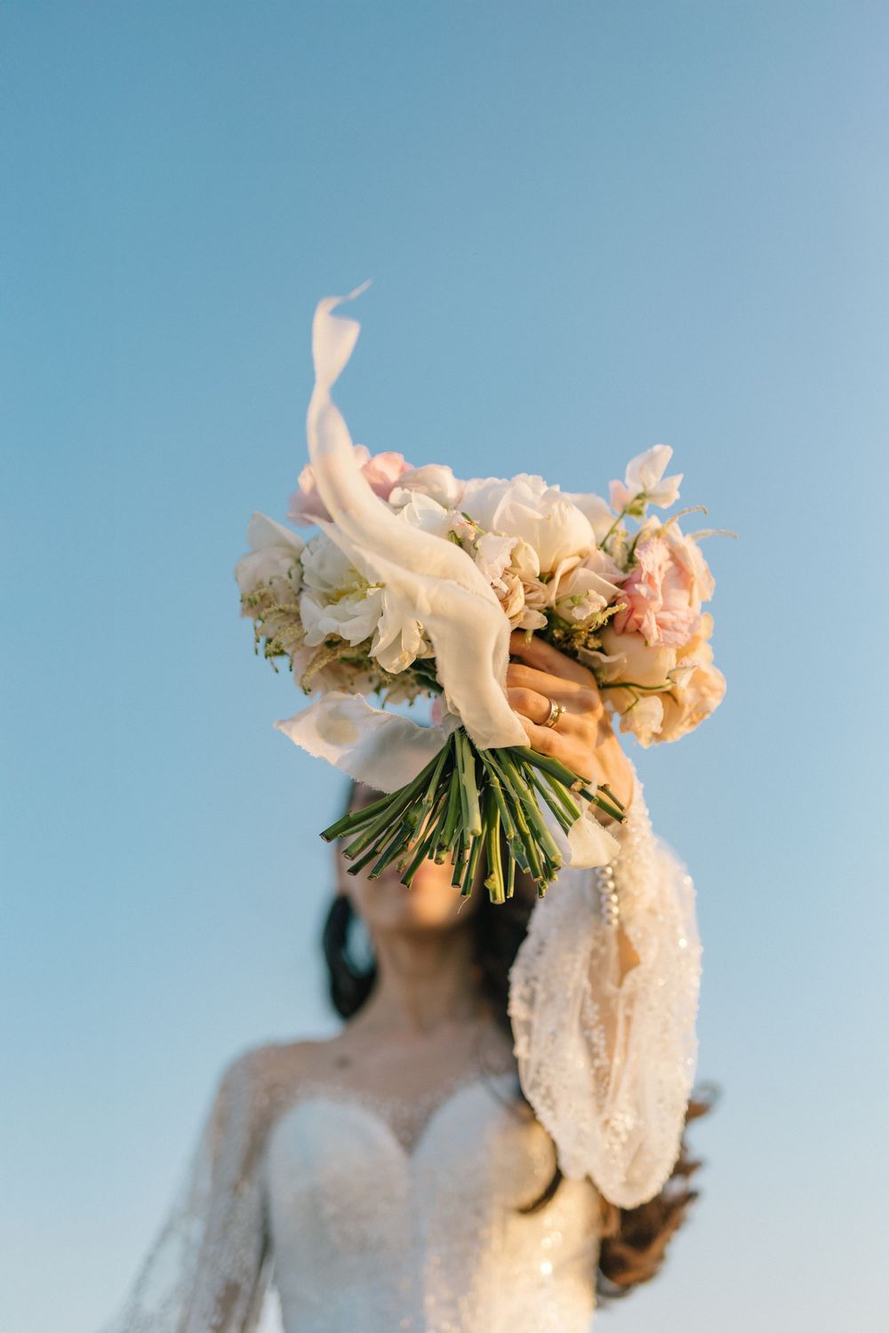 Bride in lace wedding dress holds bouquet of white and pink flowers against clear blue sky.