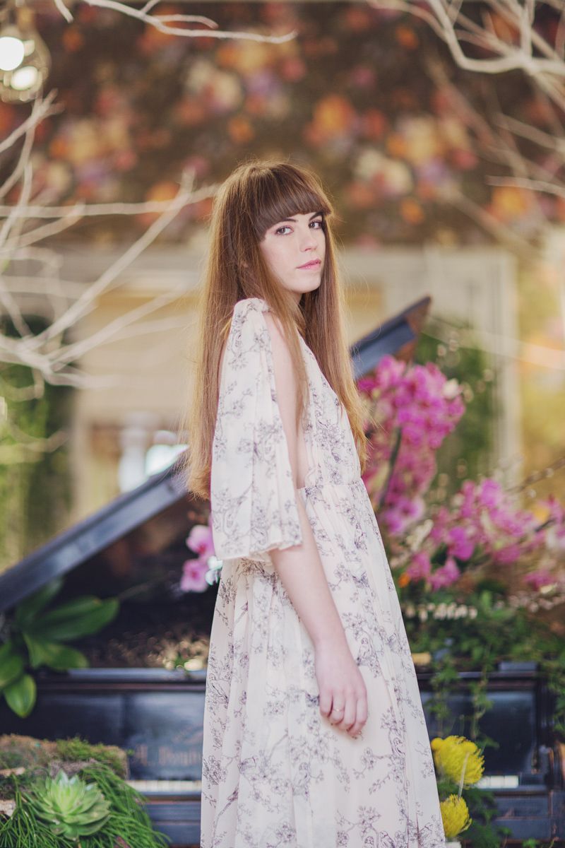 Young woman in floral maxi dress standing among colorful blooms near a vintage piano outdoors.