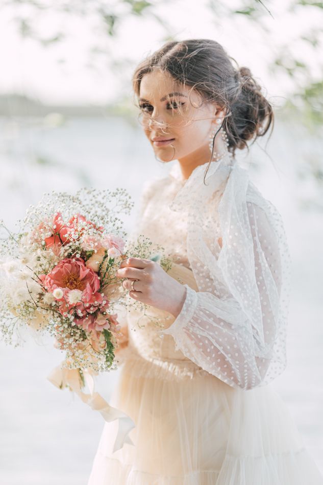 Elegant bride in lace dress holding pink floral bouquet with baby's breath, wearing veil outdoors.