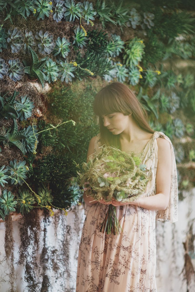 Young woman in floral dress holding green bouquet beside lush succulent wall garden.