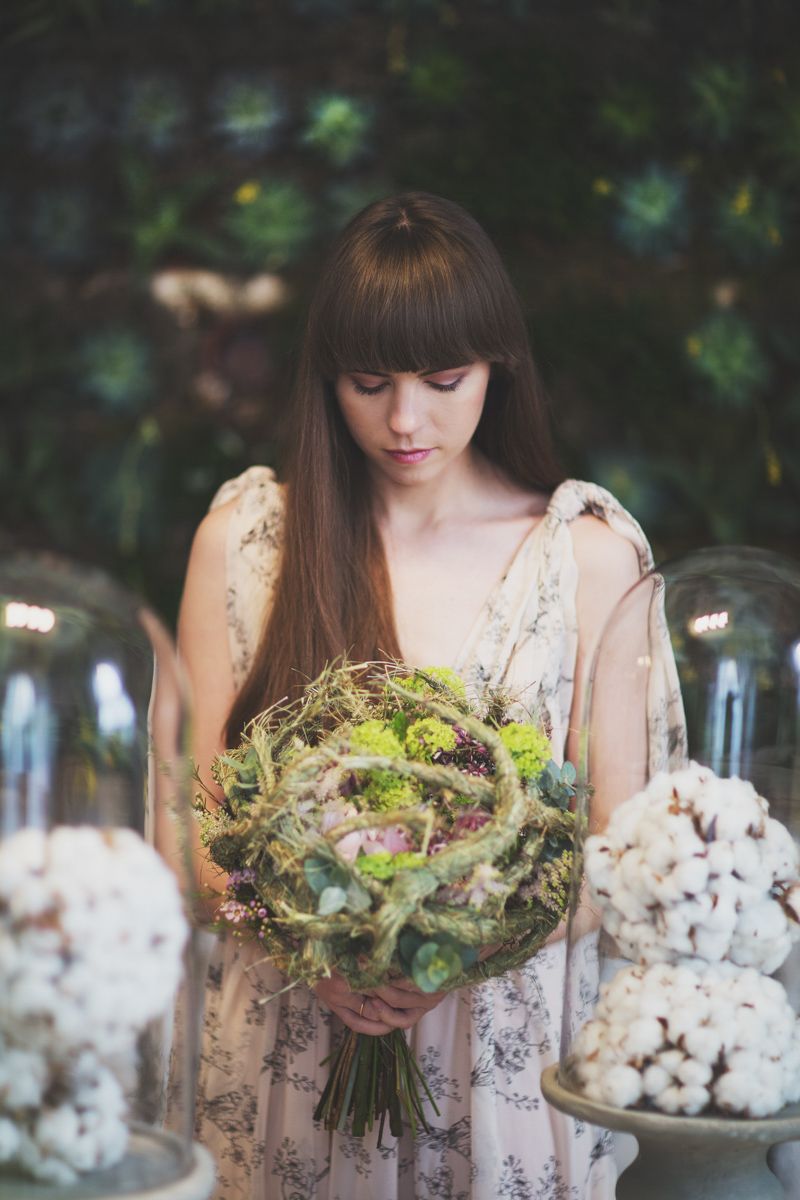 Young woman with bangs holding a lush green and pink floral bouquet, surrounded by white cotton flowers in a garden setting.