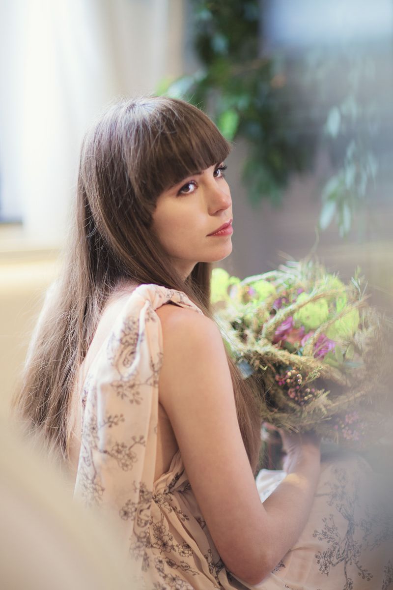 Young woman with long brown hair holding a wildflower bouquet in a floral dress, posing indoors near green plants.