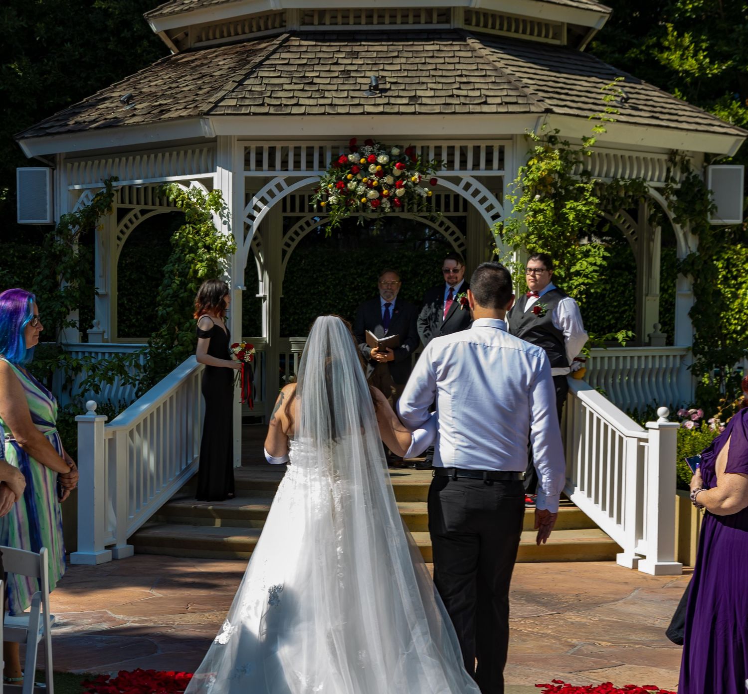 Bride and groom walking hand in hand toward a white gazebo altar decorated with red flowers at an outdoor wedding.
