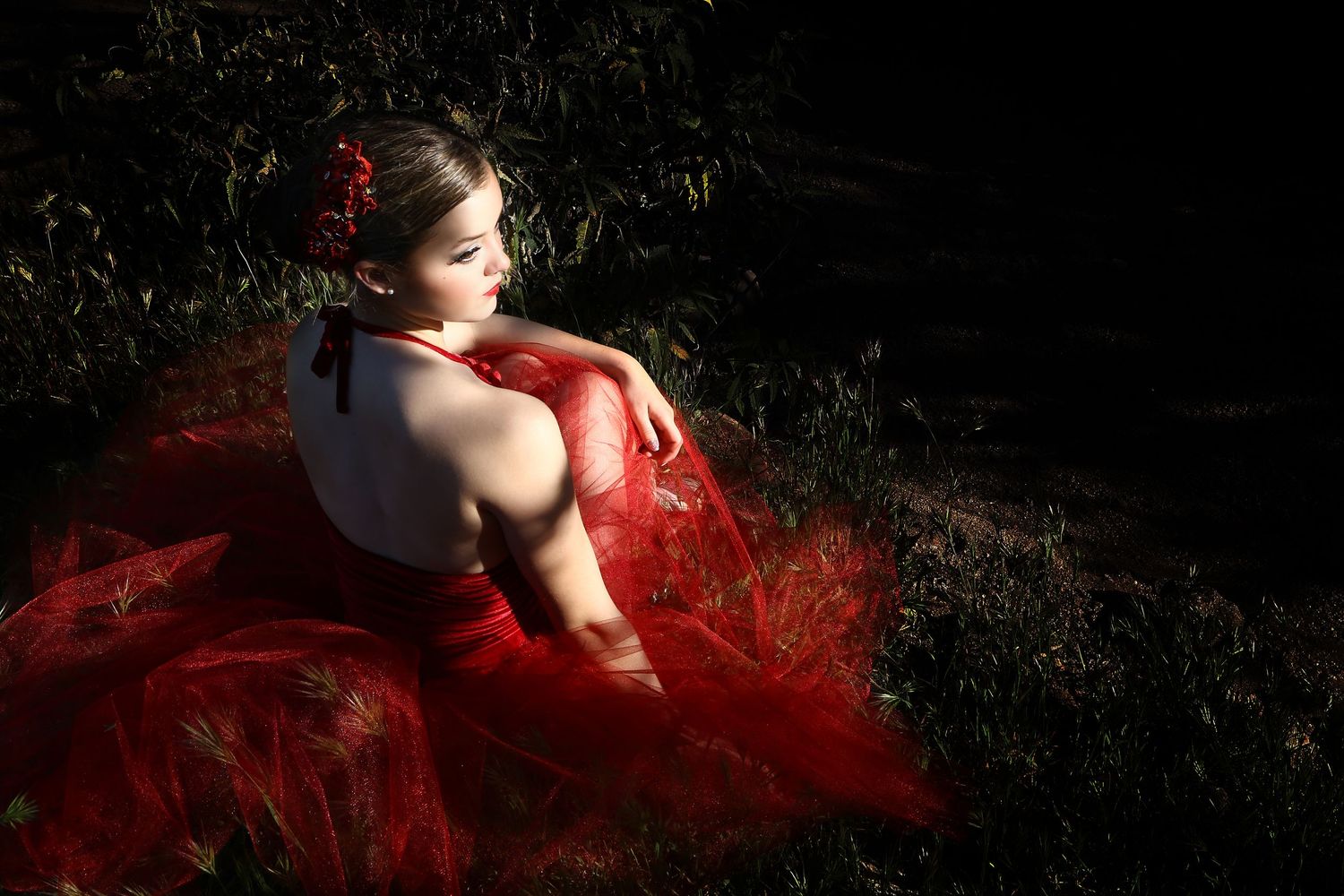 Woman in a flowing red tulle gown with a red flower in her hair sits on dark grass, her back turned toward the camera.