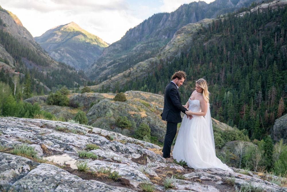 Bride and groom exchanging vows on rocky mountain summit with golden sunlit valley and forest below.