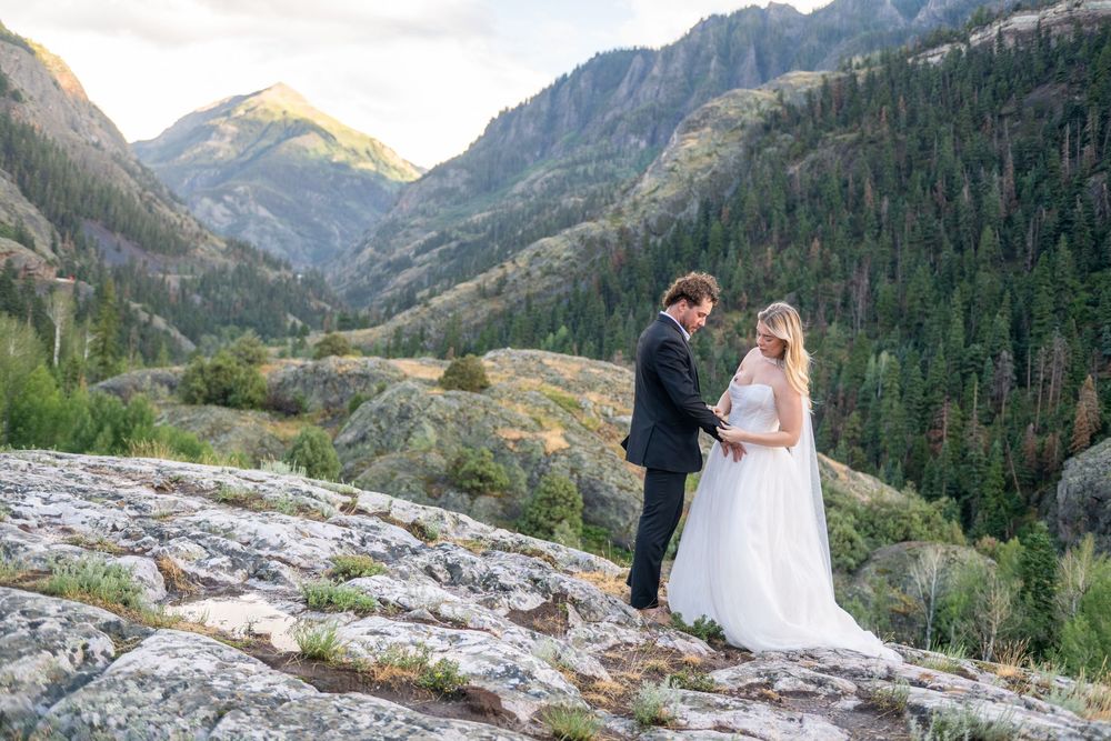 Wedding couple holding hands on rocky mountain overlook with warm sunset light illuminating the valley behind them.