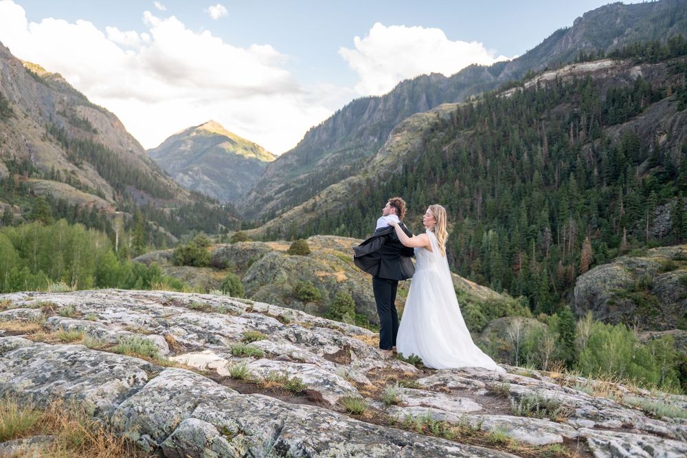 Bride and groom hold hands while standing on rocks overlooking a stunning mountain valley.