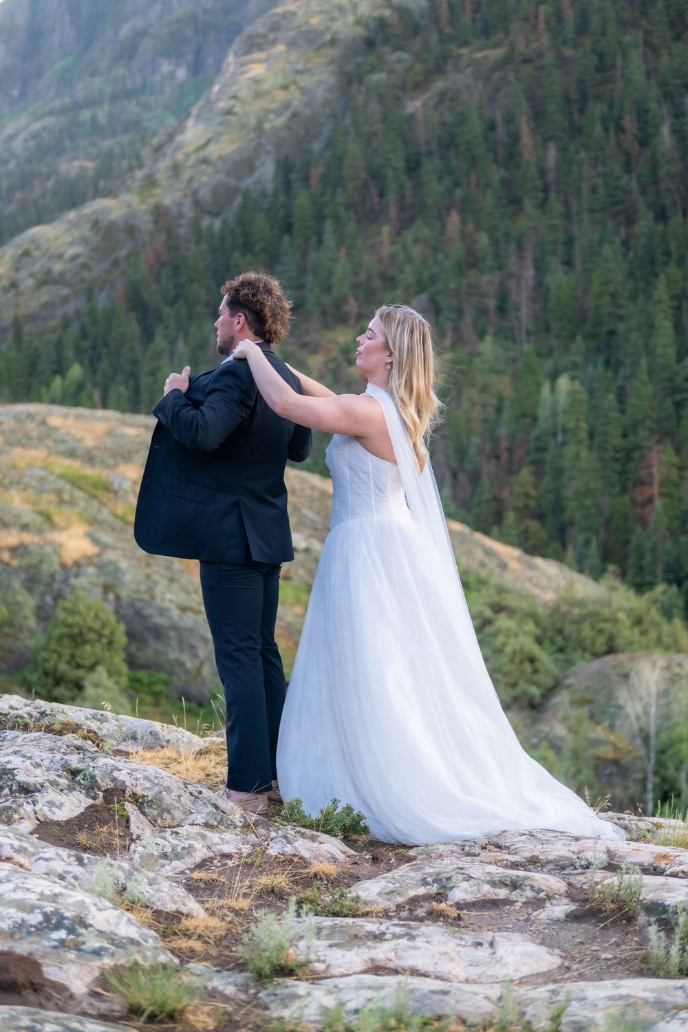 Smiling bride in white gown and groom in black suit pose on rocks with mountain forest backdrop.