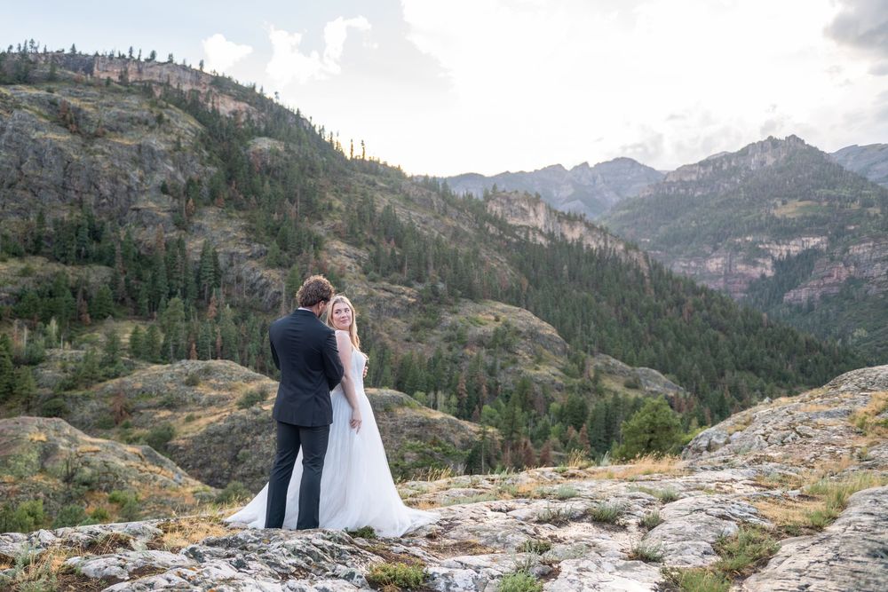 Bride and groom embrace on rocky mountain summit overlooking lush pine-covered hillsides under cloudy skies.