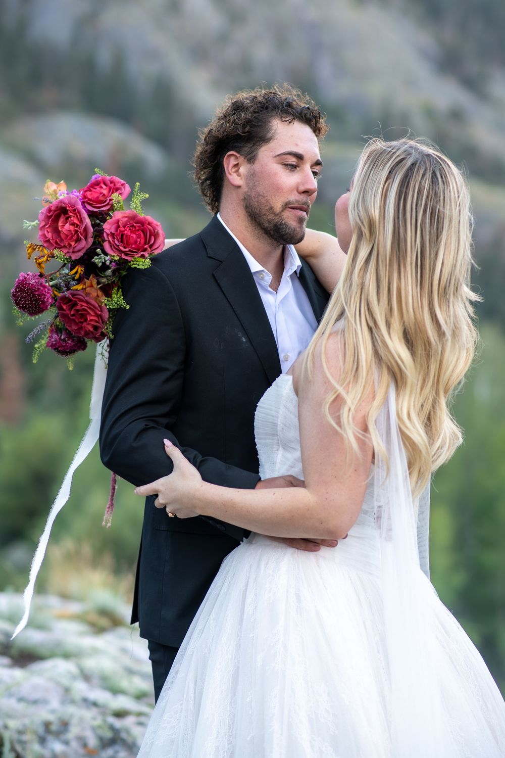 Groom embraces blonde bride holding red rose bouquet against mountain scenery.
