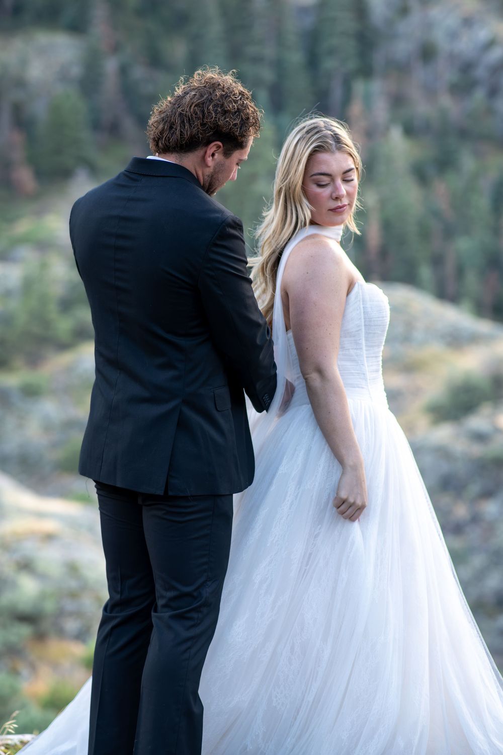 Groom in black suit holds bride's hands as she looks over shoulder in white tulle gown.