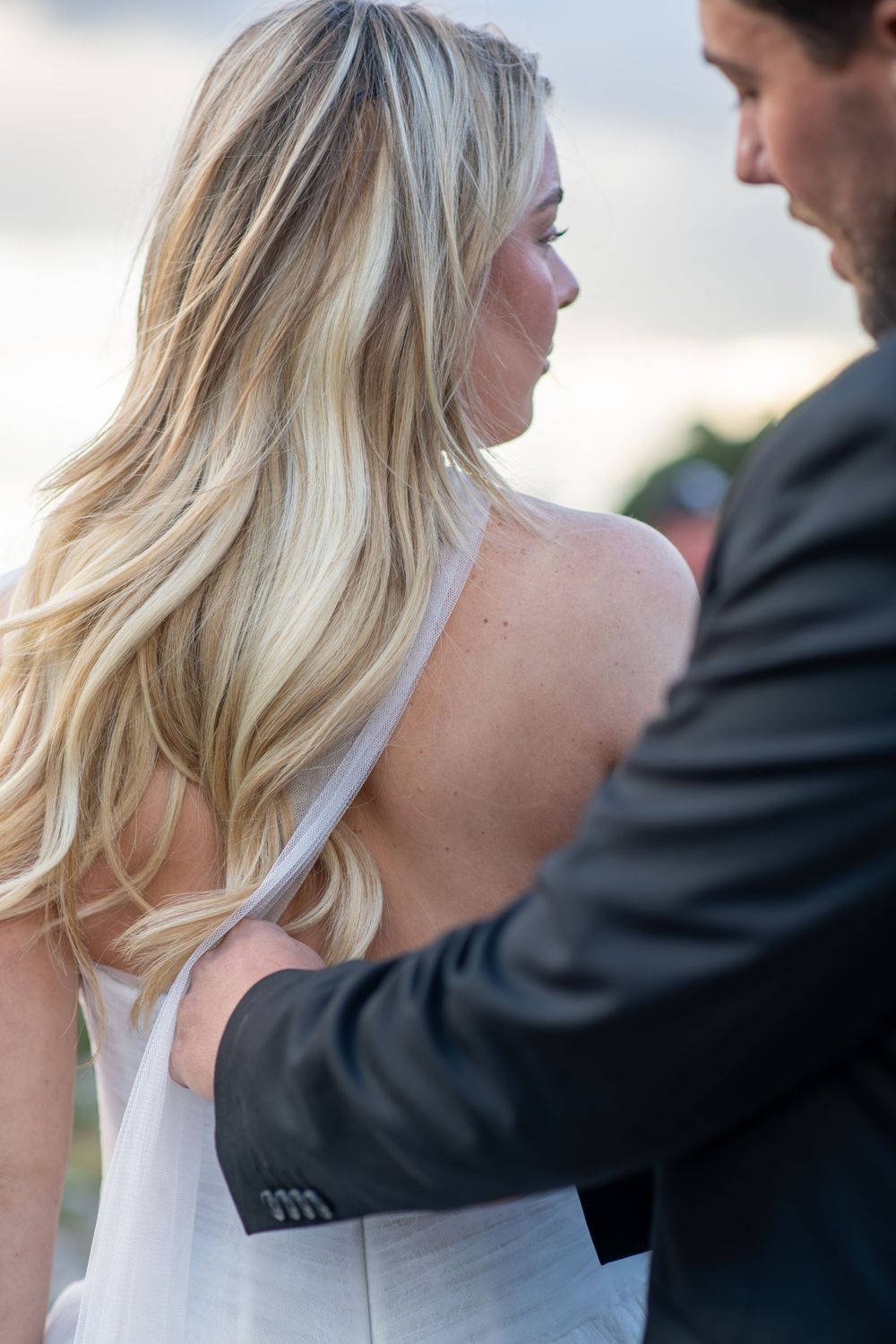 Groom in gray suit placing hand on bride's bare back during outdoor wedding ceremony.
