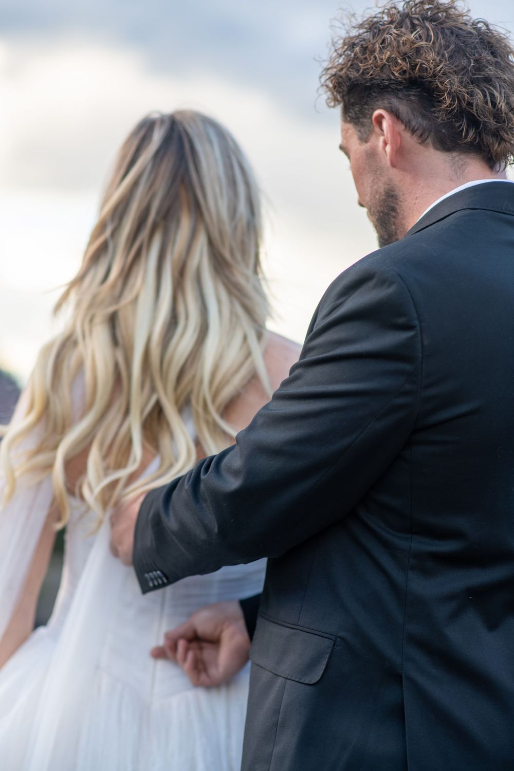 Groom in black suit holds bride's hand from behind, her blonde waves flowing in the breeze.
