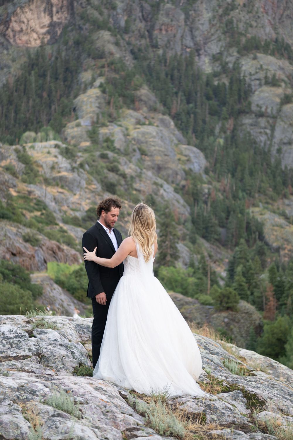 Bride and groom embrace on rocky mountain terrain surrounded by lush green forest.