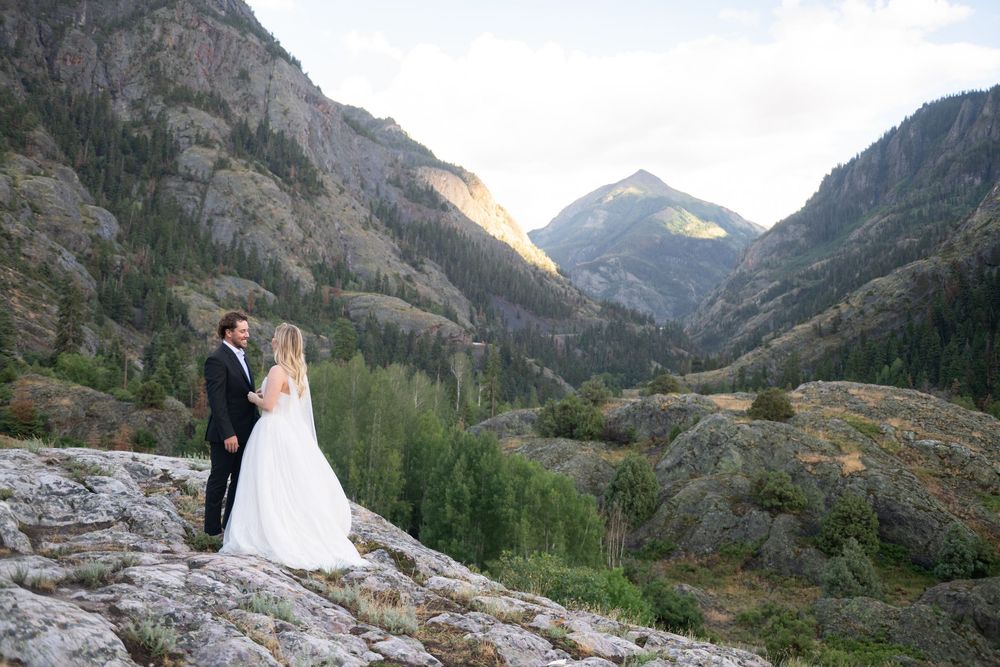 Wedding couple stands hand in hand on rocks overlooking a sunlit mountain valley.