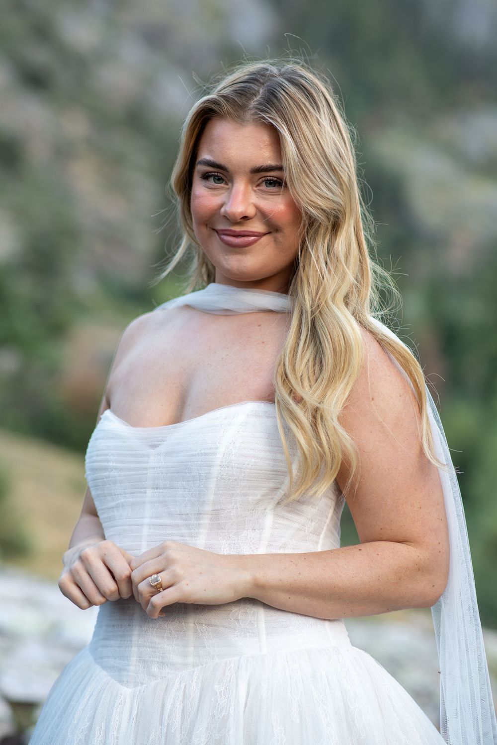 Beautiful blonde bride in strapless white tulle gown smiling softly with mountain scenery in background.