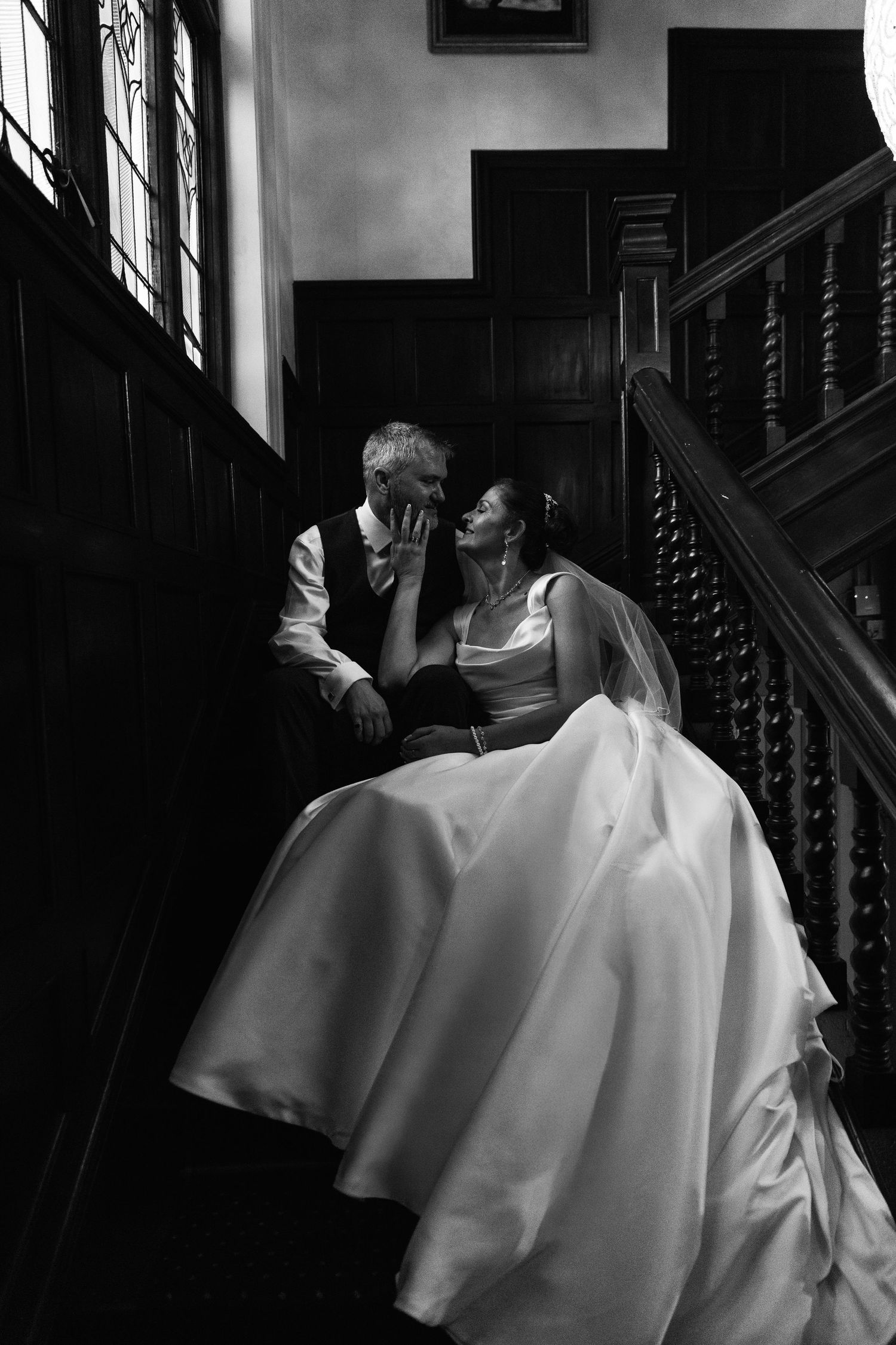 Black and white photo of a bride and groom sharing an intimate moment on a grand staircase in a historic venue.