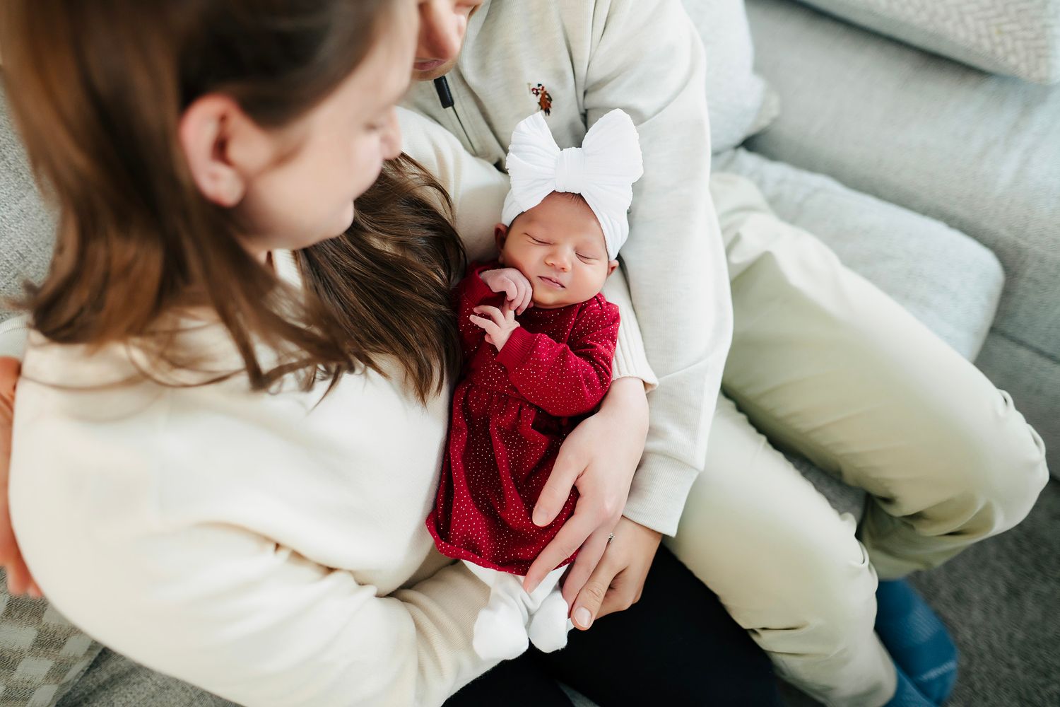 Parents cradling a sleeping newborn baby in a red outfit with a white bow headband on a gray couch.