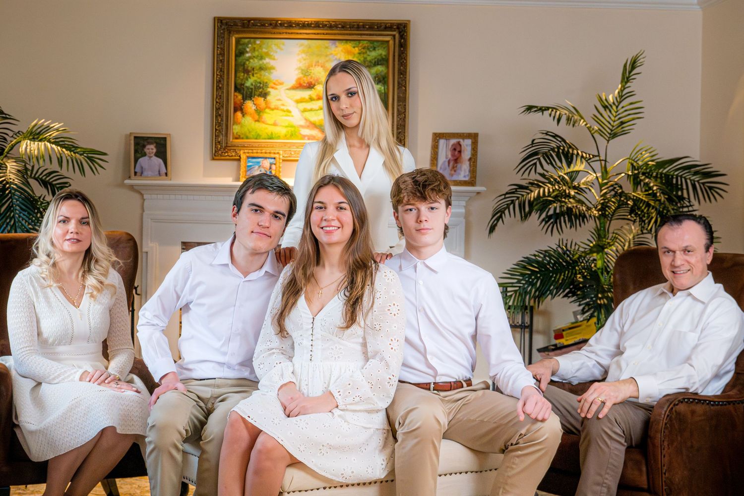 A smiling family of six poses together in a living room, all wearing white and beige outfits for a formal portrait.