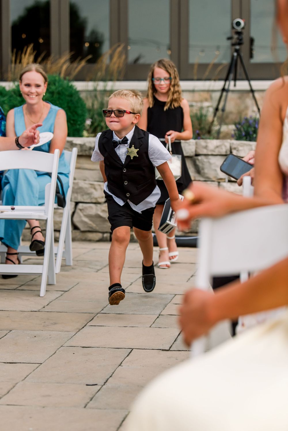 Young ring bearer in sunglasses and vest runs down the aisle at an outdoor wedding ceremony.