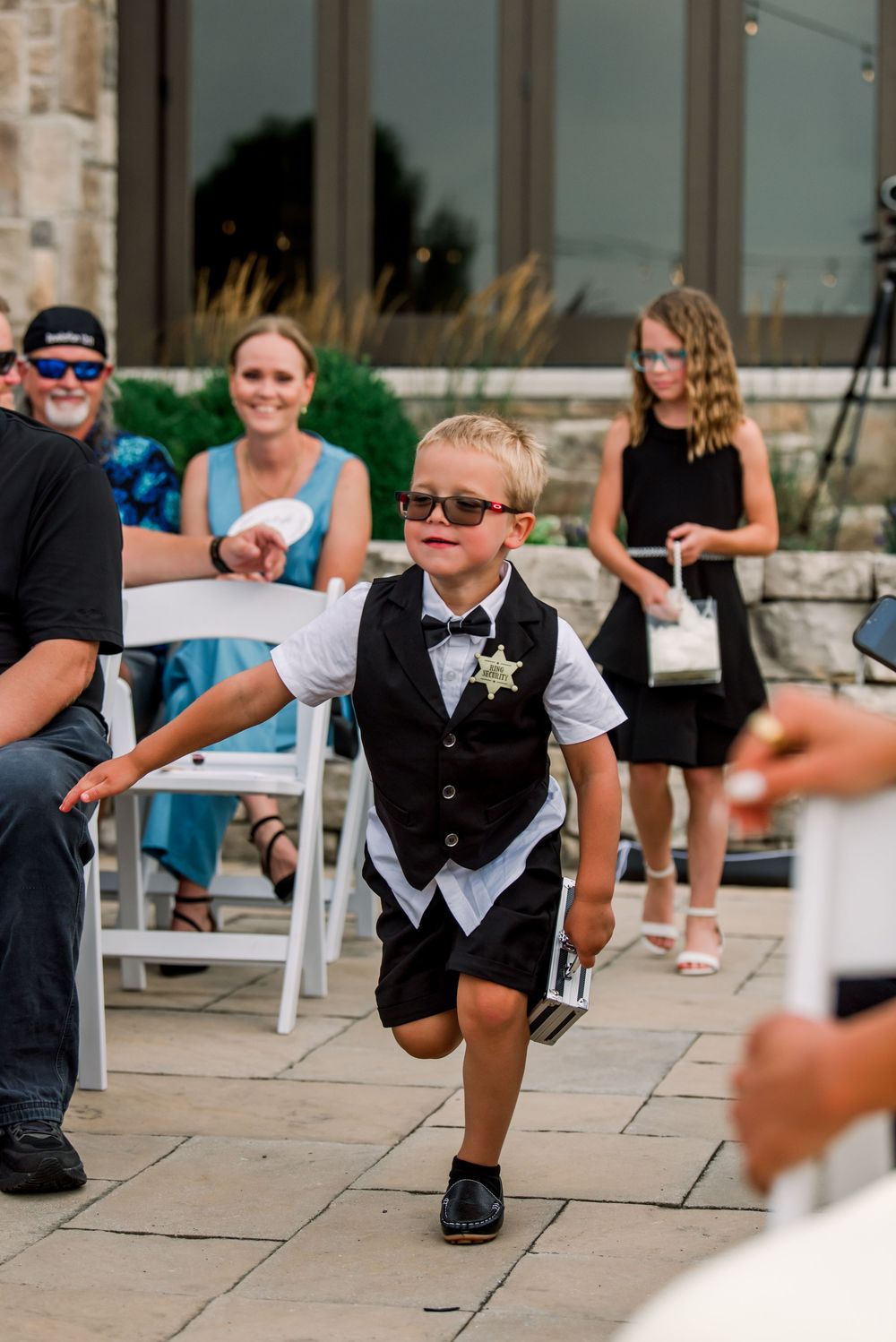 Young ring bearer in black vest, bow tie, star badge, and sunglasses strutting down wedding aisle with confidence.