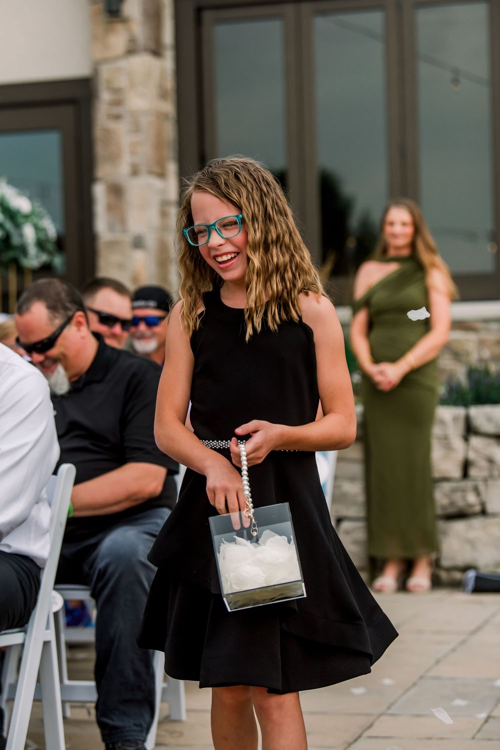 Young girl in black dress with glasses smiles while carrying a flower girl basket at an outdoor wedding ceremony.