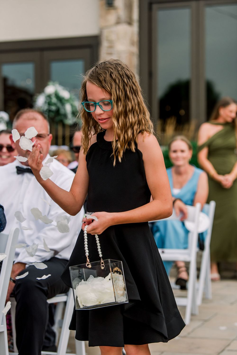 Young flower girl in black dress with teal glasses carries clear purse with white petals at outdoor wedding ceremony.
