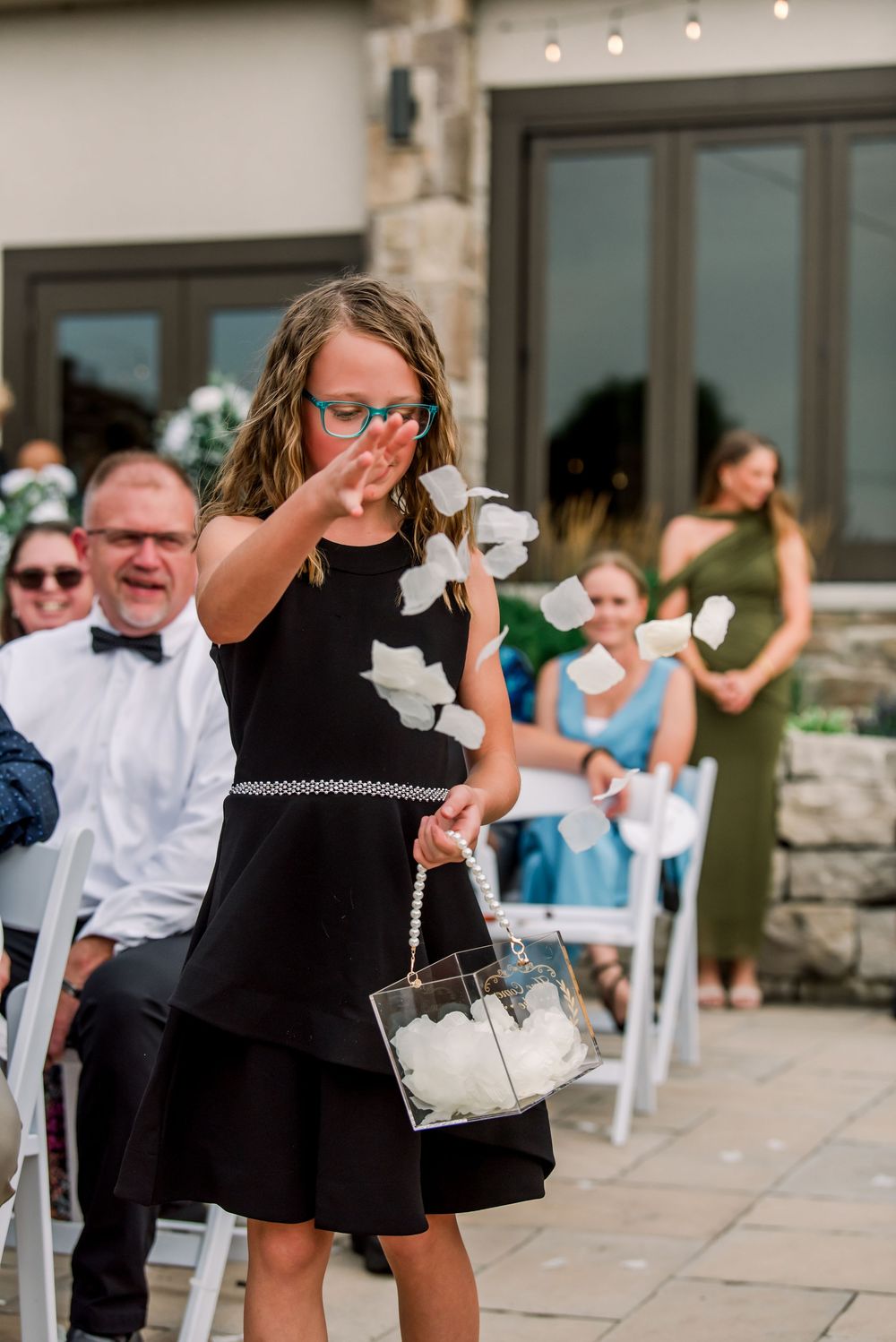 Young flower girl in black dress tossing white petals at an outdoor wedding ceremony with guests seated behind her.