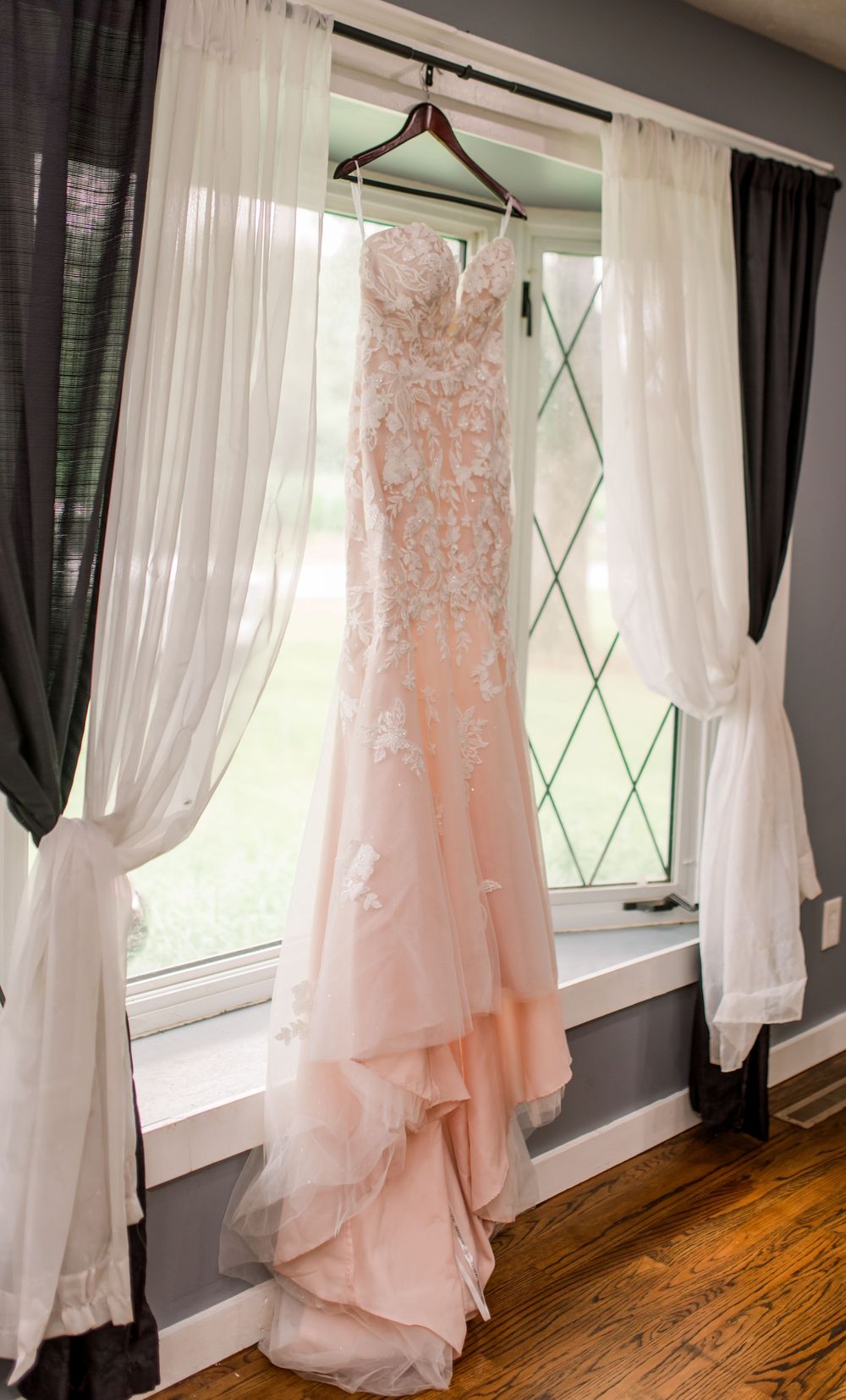 Blush pink lace mermaid wedding dress hanging in a sunlit window with sheer white curtains and diamond-pane glass.
