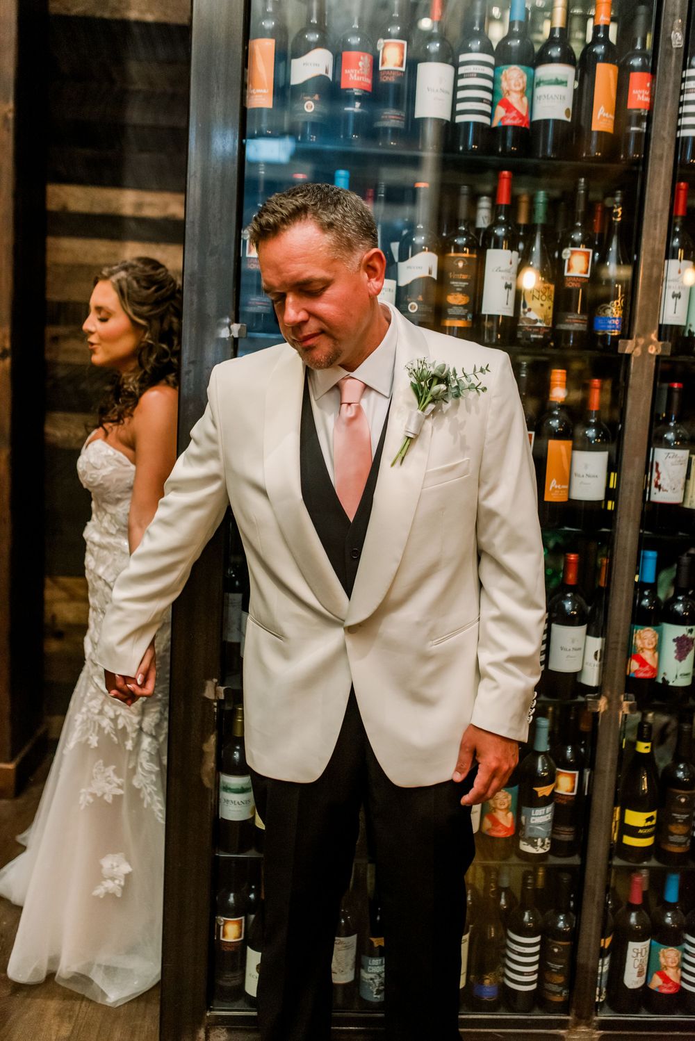 Groom in white tuxedo jacket with pink tie stands by wine cellar as bride in lace gown looks on during wedding.