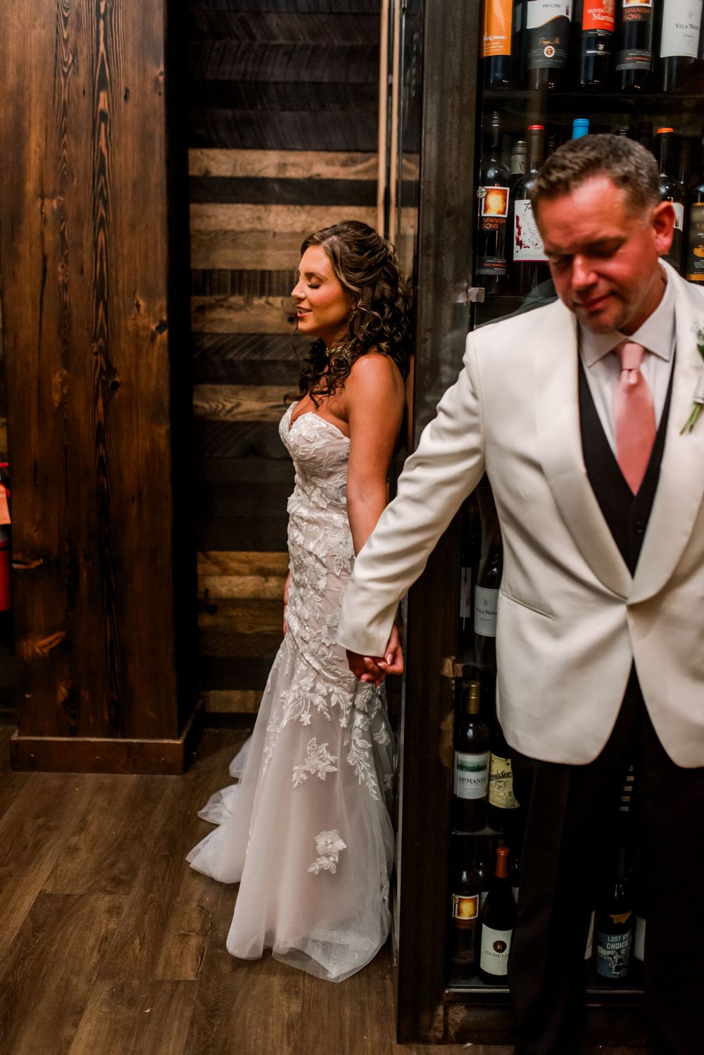 Bride in lace wedding gown holds groom's hand in white tuxedo jacket near rustic wood wine cellar.