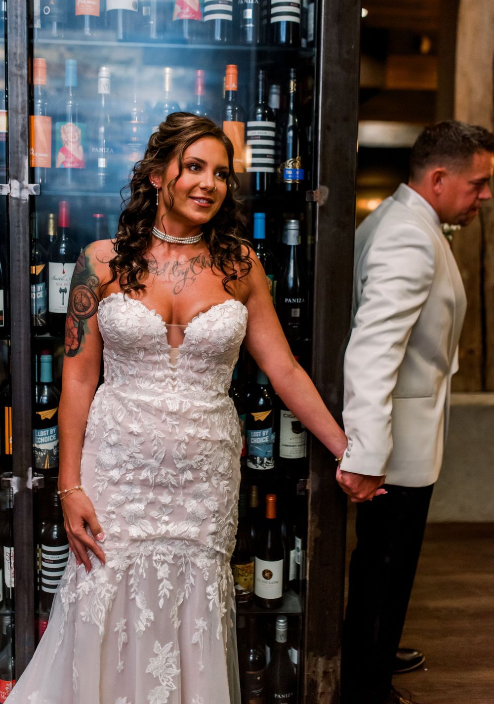 Bride in lace mermaid wedding dress stands by wine cellar holding hands with groom in white tuxedo jacket.
