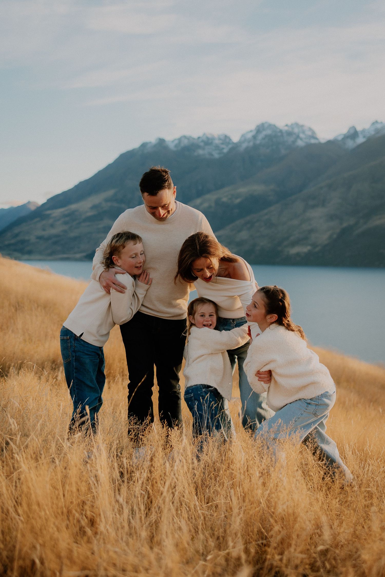 Family of five laughing and hugging in golden grass field with mountain lake backdrop in New Zealand.