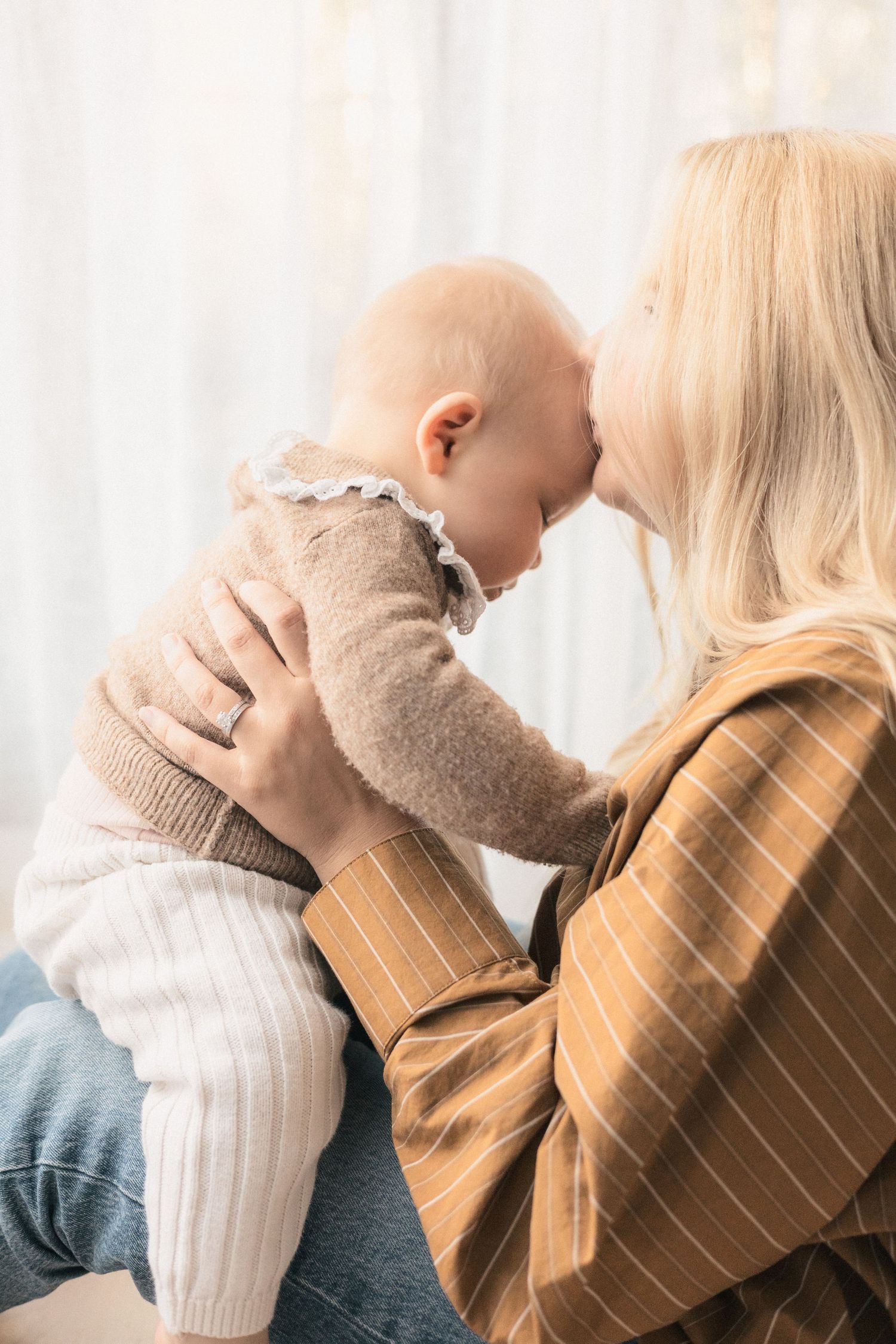 Blonde mother tenderly touching foreheads with her smiling baby, sharing a loving moment indoors.