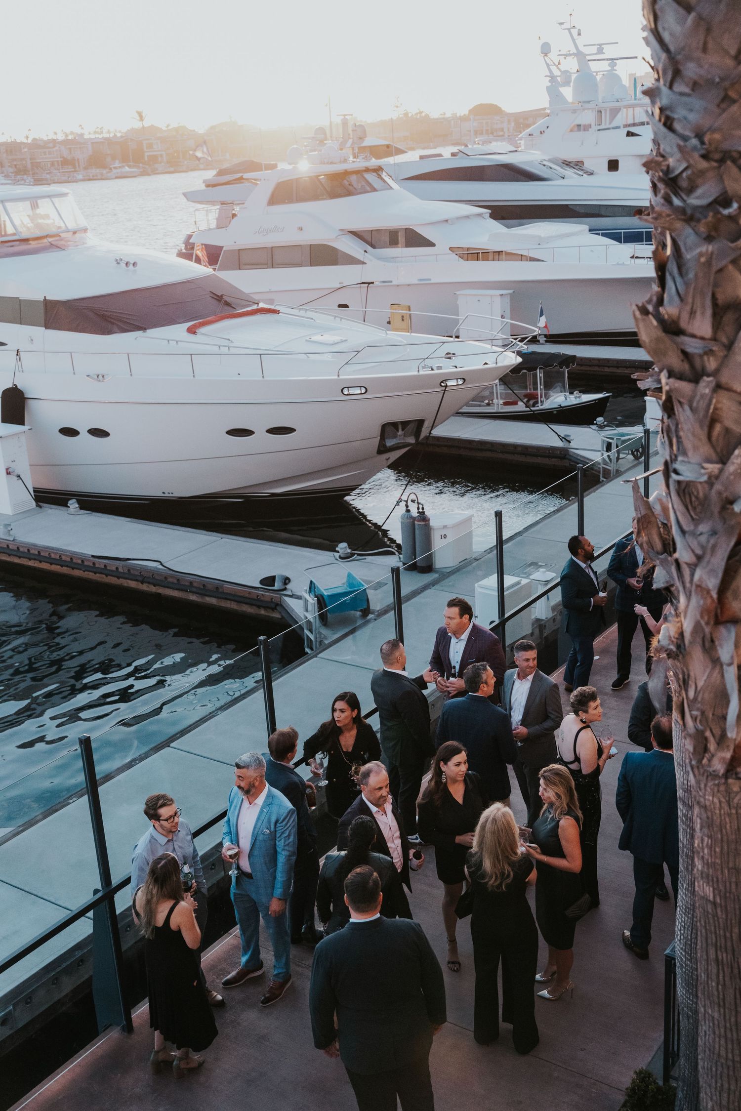 Elegantly dressed guests mingle on a marina dock at sunset, surrounded by luxury yachts at an upscale outdoor event.