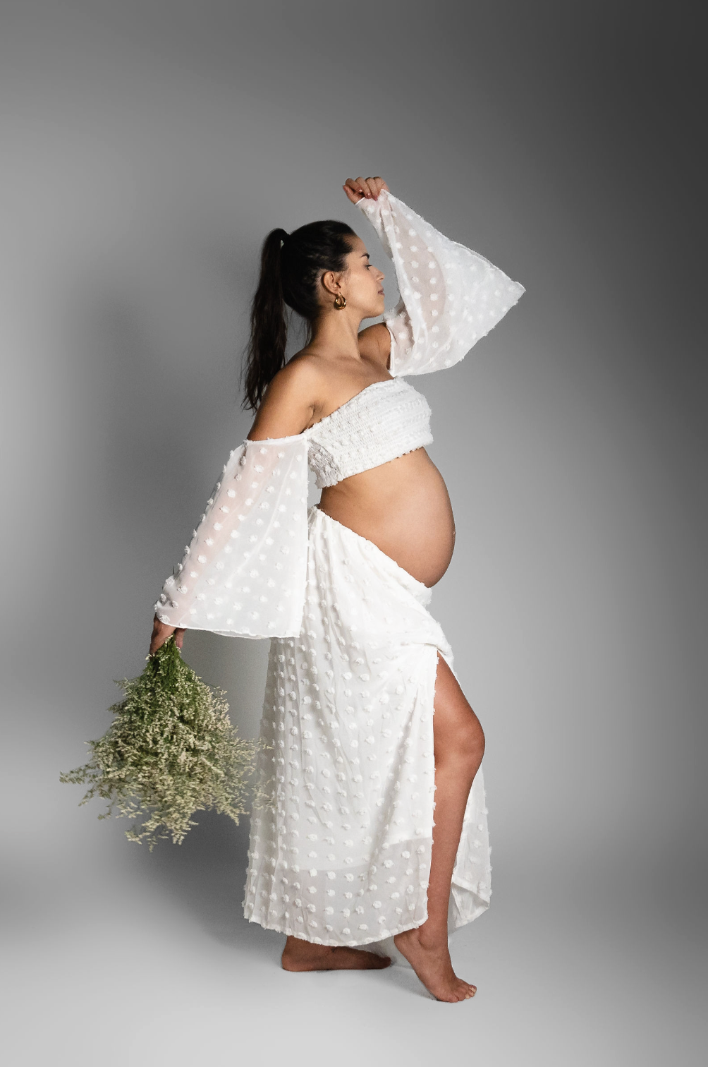 Pregnant woman in white polka dot off-shoulder dress holding wildflowers, posing gracefully on gray background.