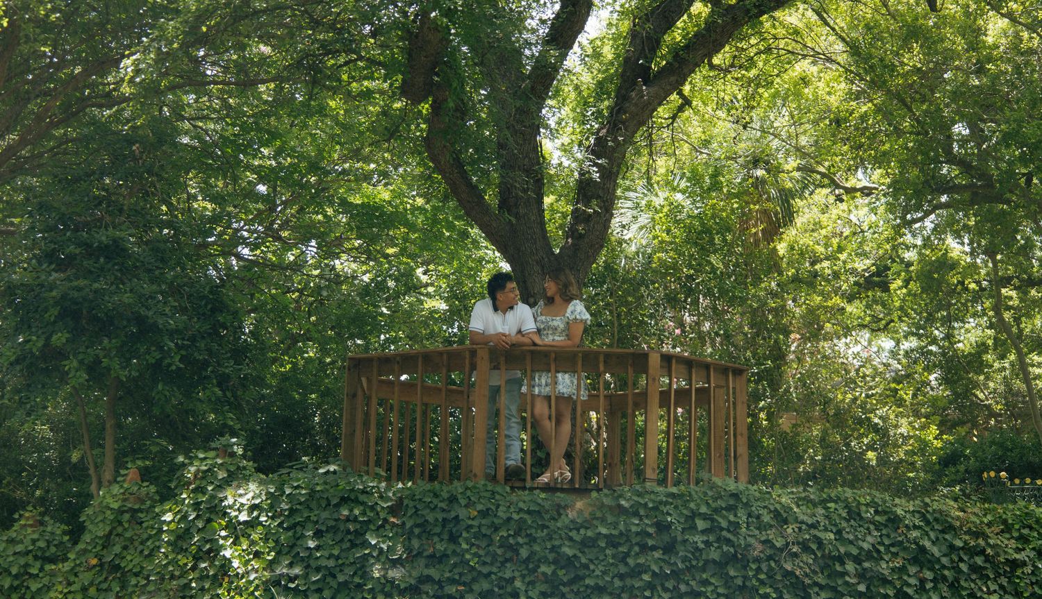 Two people relaxing in a wooden hexagonal treehouse platform surrounded by lush green trees and ivy.