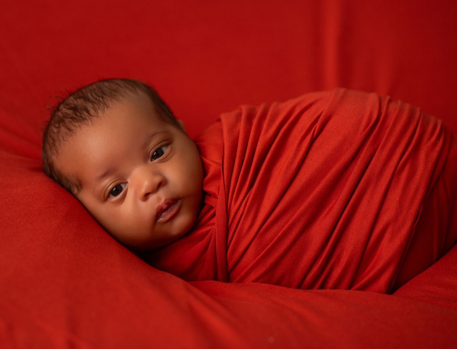 Newborn baby swaddled in red fabric on a red background, peacefully resting in a cozy wrap.