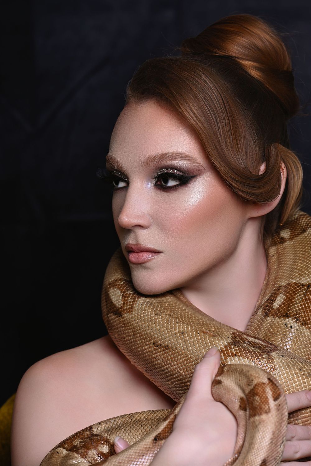 Young woman with dramatic smoky eye makeup and auburn hair posing with a large python snake draped around her shoulders.