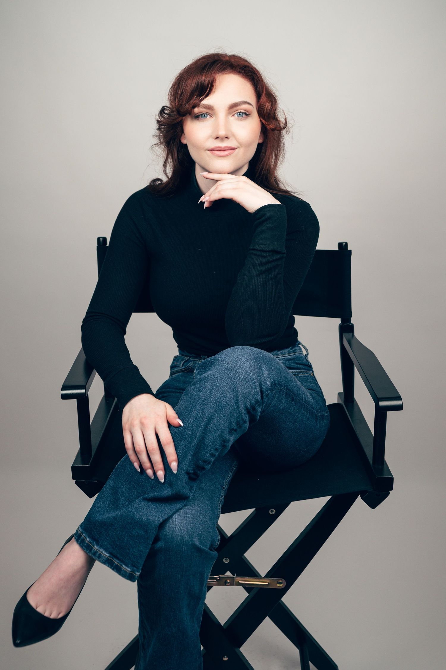 Woman in black turtleneck and jeans sitting relaxed in a director's chair against a gray background.