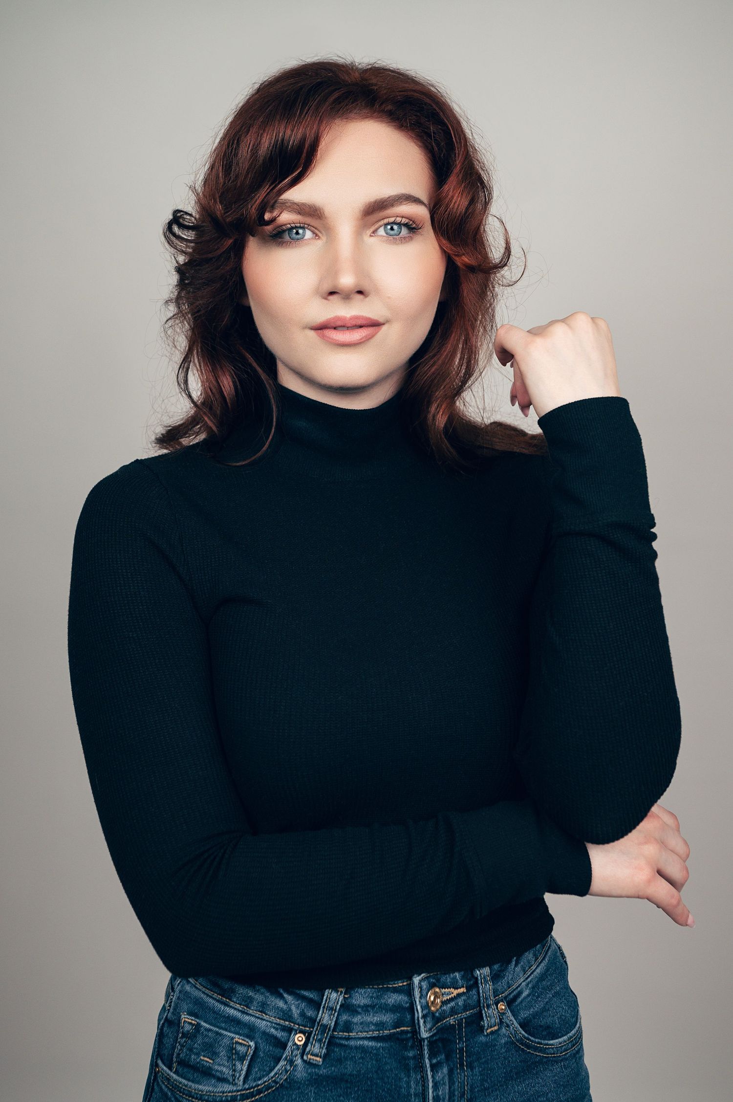 Young woman with curly auburn hair in black turtleneck and jeans poses confidently against gray background.