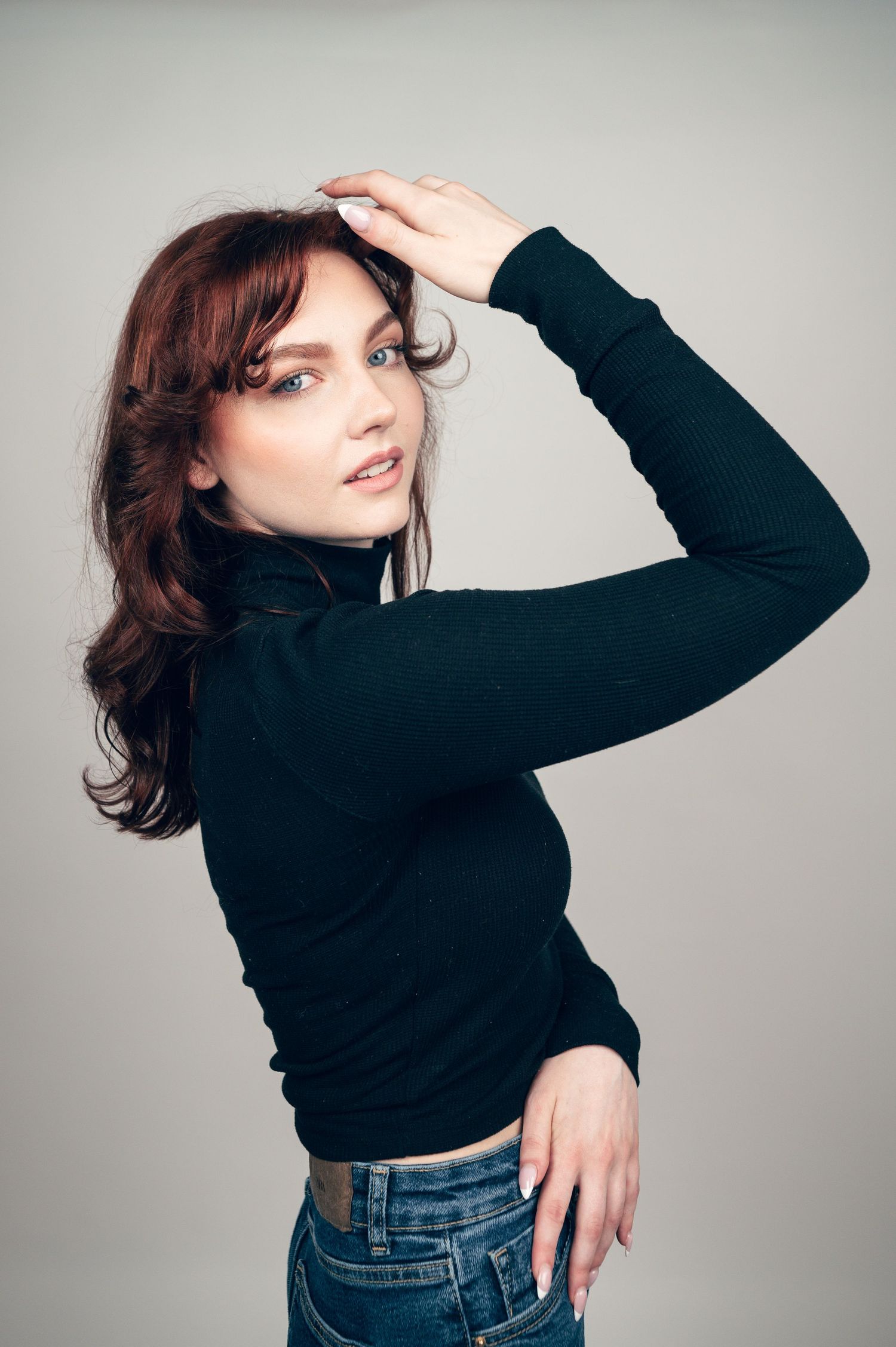 Young woman with brown hair in black turtleneck and jeans poses confidently against gray background.