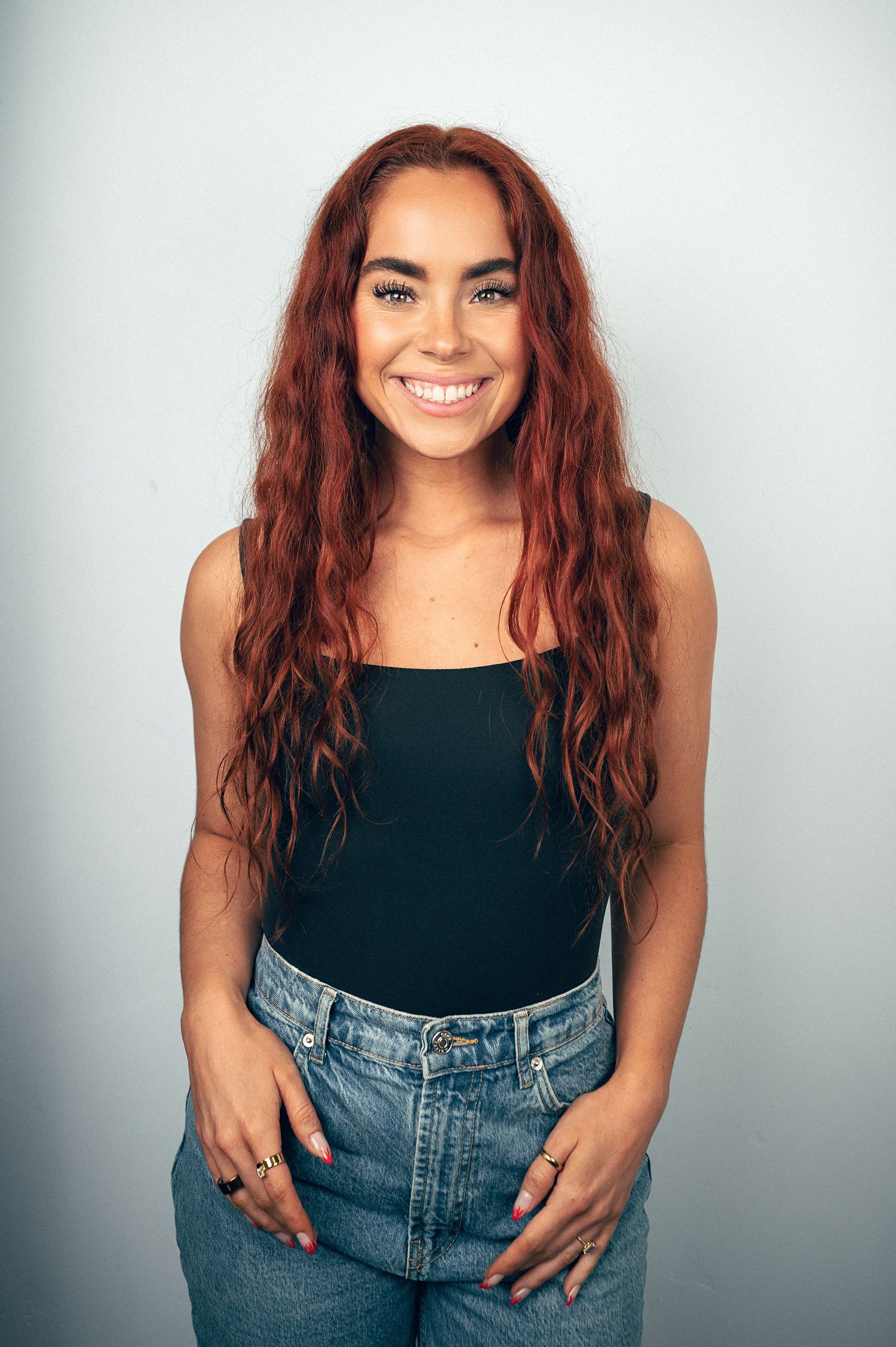 Young woman with long red wavy hair smiling, wearing black tank top and blue jeans against light background.