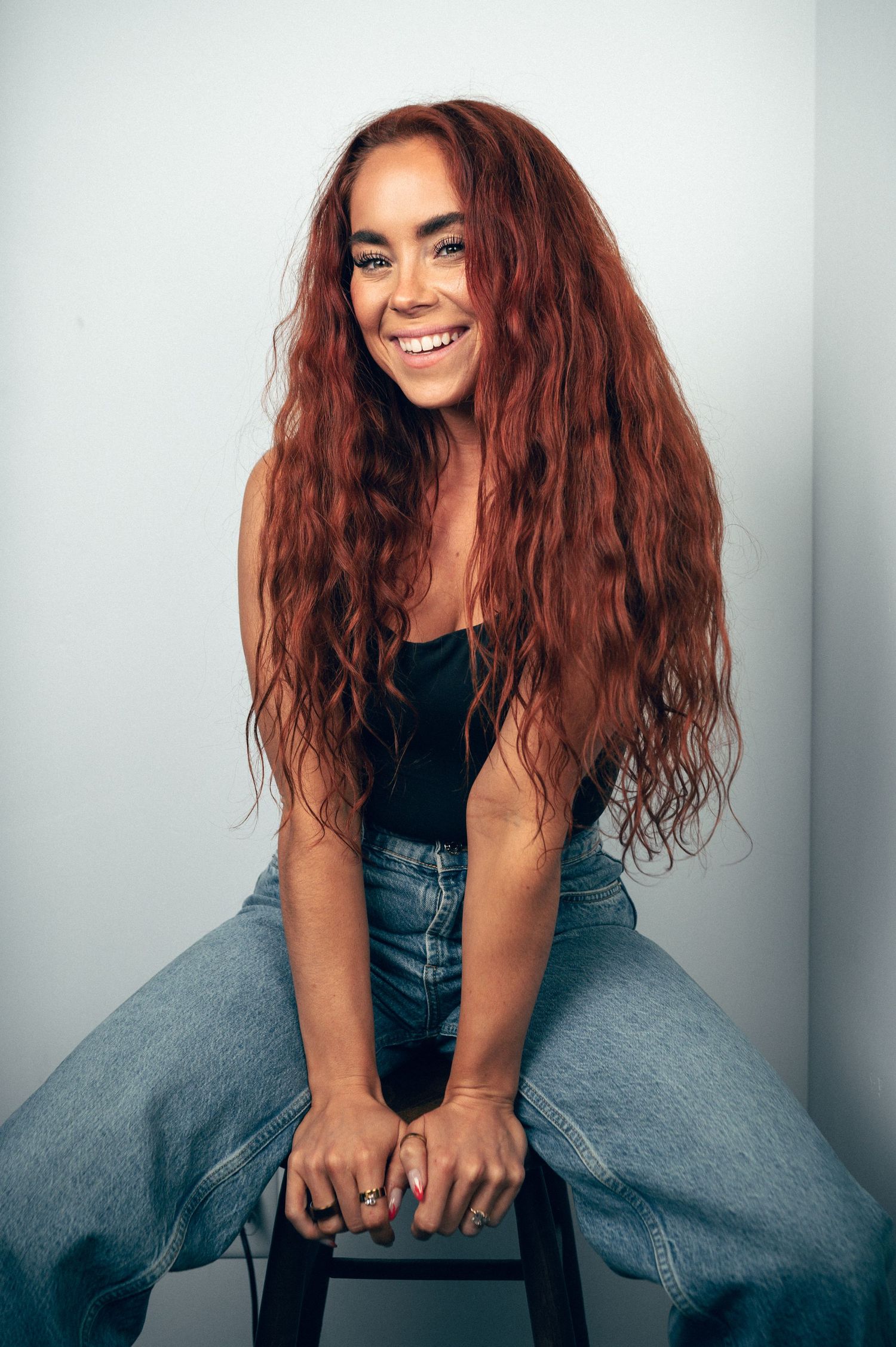 Young woman with long wavy red hair smiling, wearing black top and jeans, seated on stool against light background.