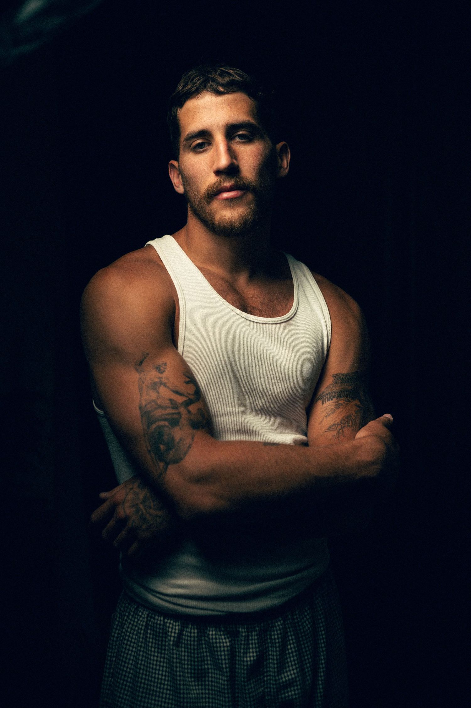 Muscular man with tattoos in white tank top, arms crossed, dark background studio portrait.
