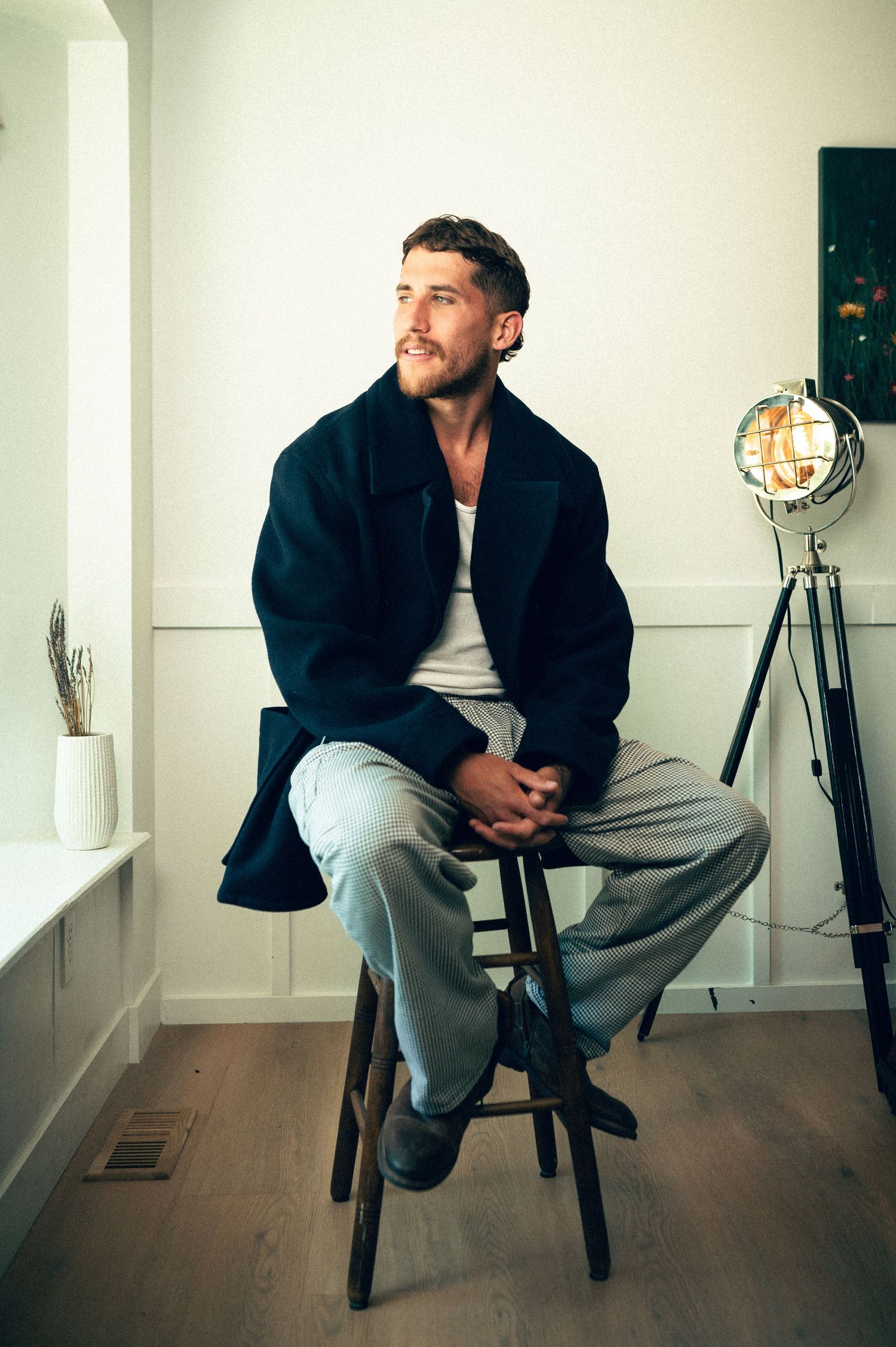 Man in dark jacket and grey pants sitting on wooden stool in minimalist white room with industrial lamp.