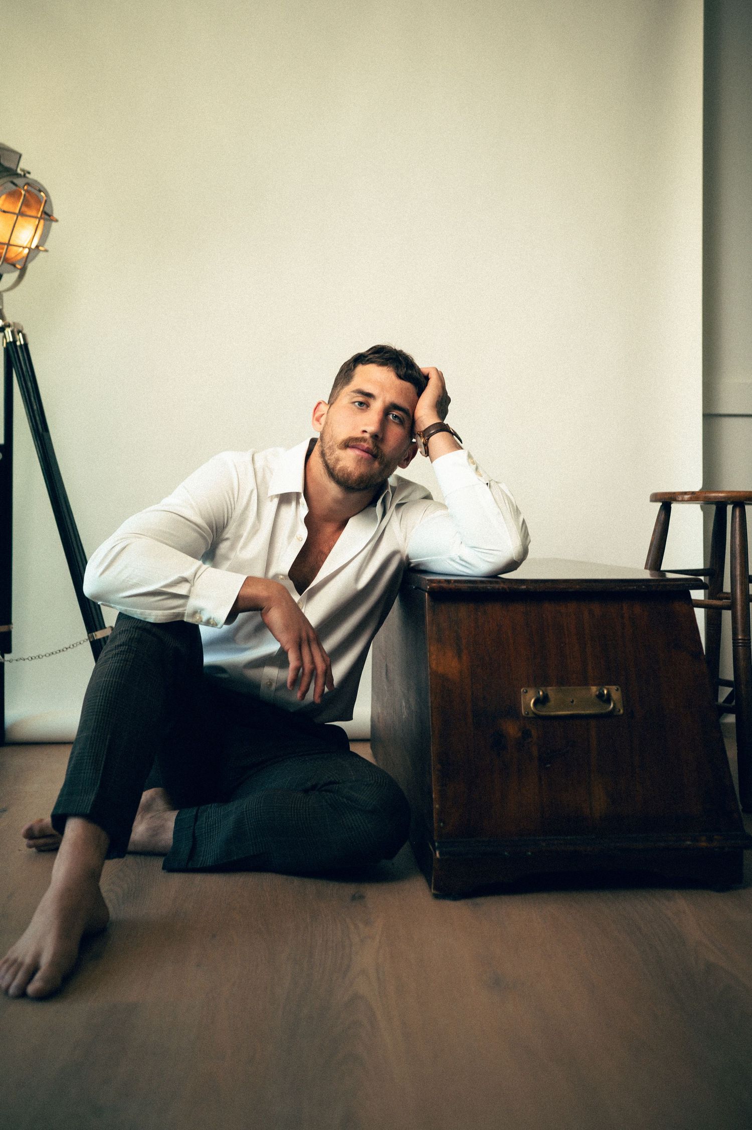 Man in white shirt and black pants sitting on floor leaning against wooden chest beside vintage floor lamp.