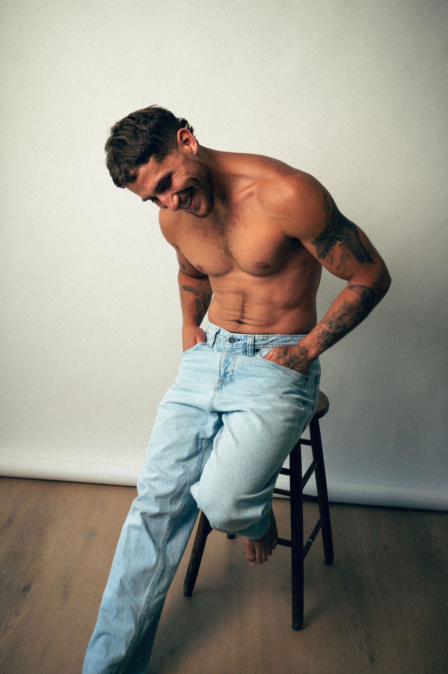 Shirtless tattooed man in light blue jeans sitting on a stool against a white wall.