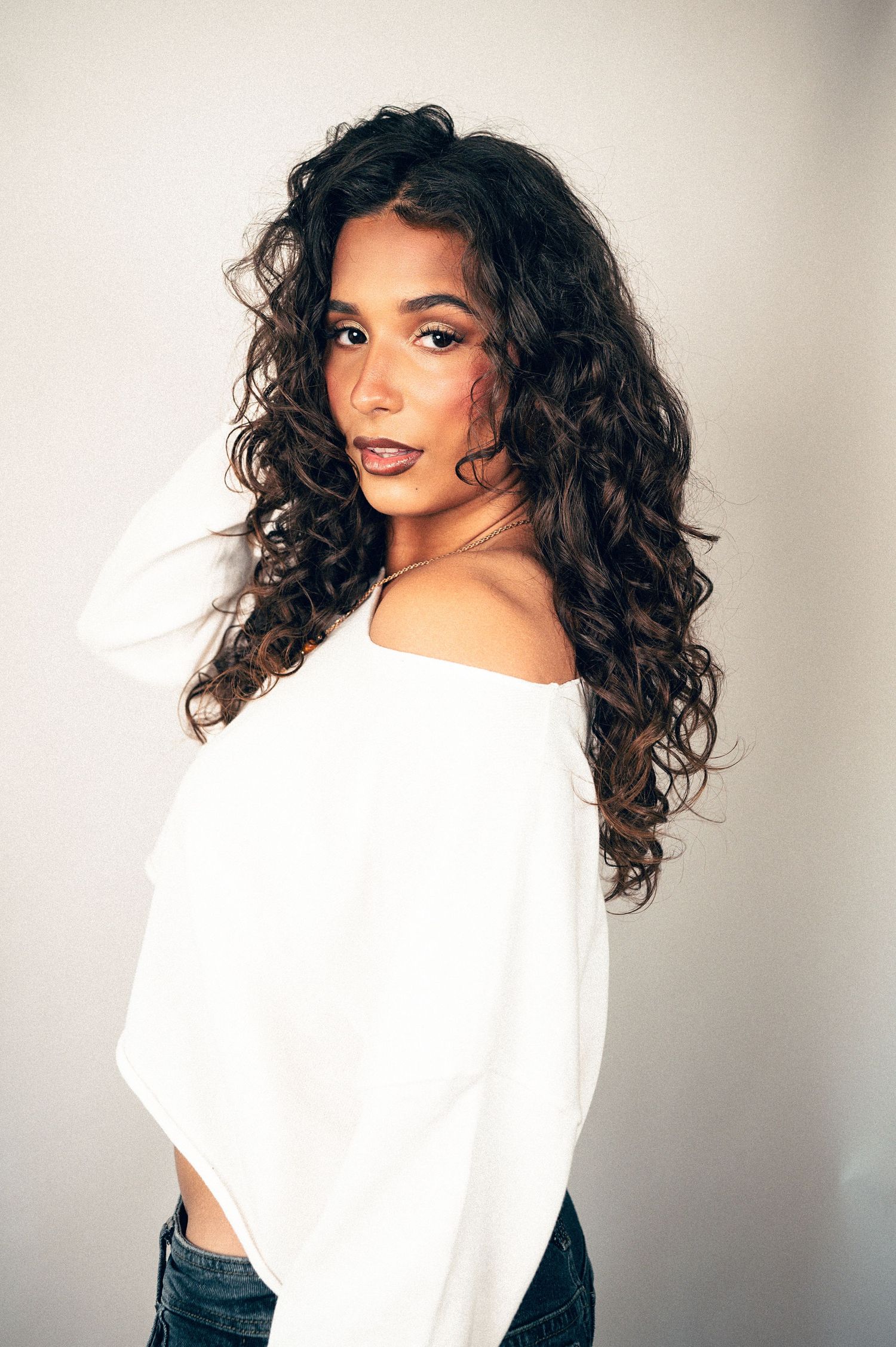 Young woman with long curly dark hair poses in white off-shoulder top and jeans against light background.