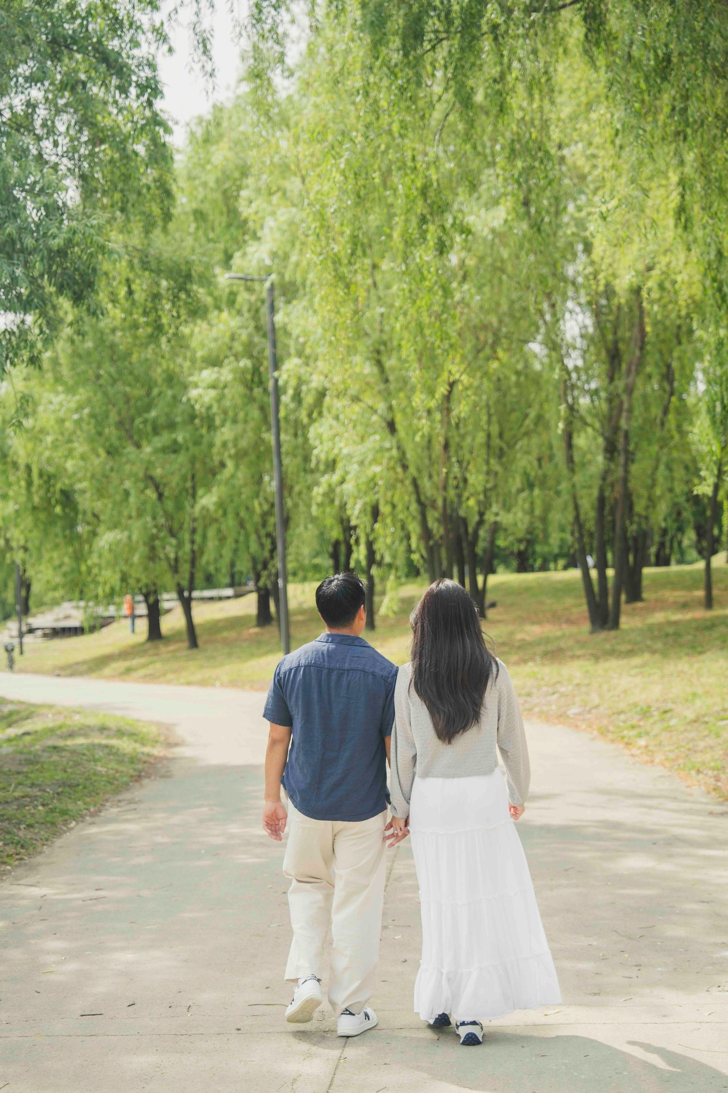 Couple holding hands walking along a tree-lined park path, viewed from behind, woman in white skirt.